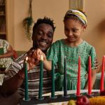 A man and a girl light candles on a kinara for Kwanzaa, with two women sitting in the background in a living room. - PsychDiscouse