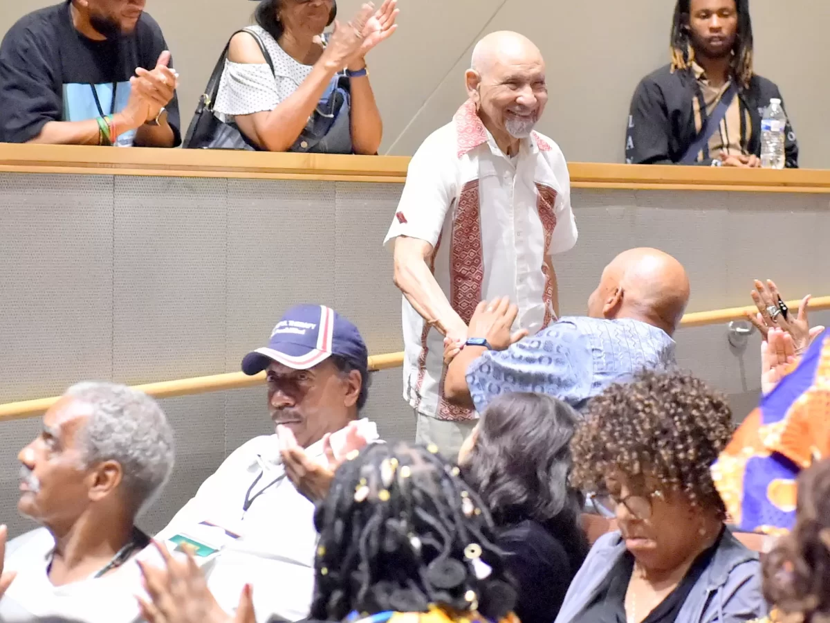 A man stands and smiles as people around him applaud in a crowded auditorium; some in balcony seats also clap and watch the scene below. - PsychDiscouse