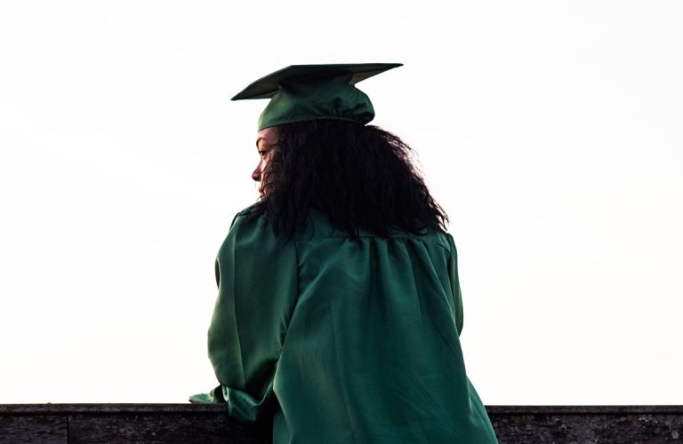 Person in a green graduation cap and gown stands outdoors, facing away from the camera, looking into the distance. - PsychDiscouse