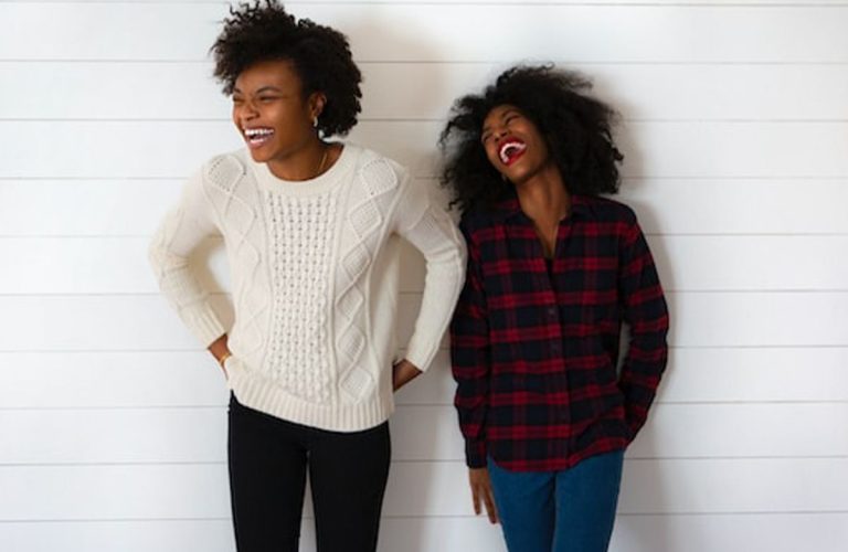 Two women standing against a white shiplap wall, smiling and laughing. One wears a white sweater and black pants, the other a red plaid shirt and blue jeans. - PsychDiscouse