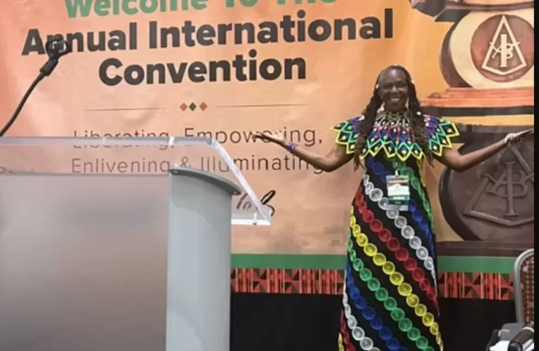 A woman in a colorful dress stands on stage, smiling, in front of a banner for The Association of Black Psychologists’ Annual International Convention. - PsychDiscouse