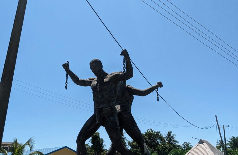 Bronze sculpture of two figures holding broken chains, standing on a raised platform outdoors, with buildings, trees, and a clear blue sky in the background. - PsychDiscouse