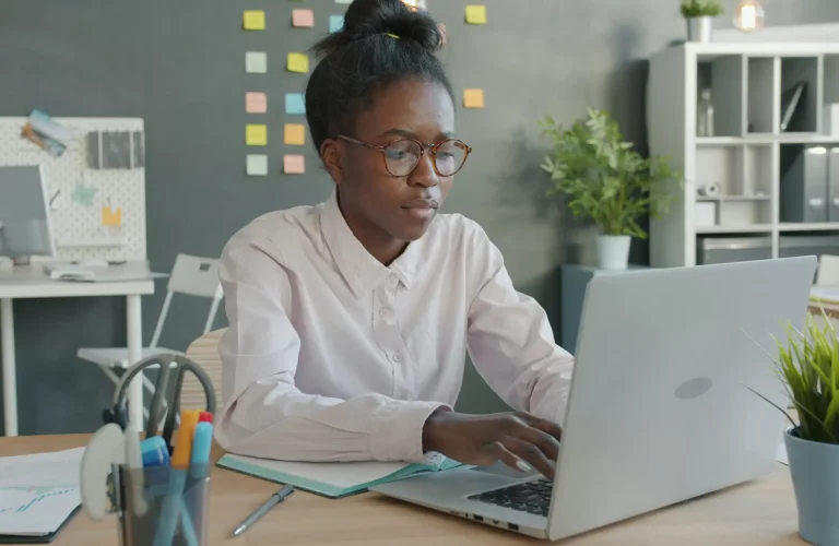 Person wearing glasses works on a laptop at a desk in a modern office with sticky notes on the wall and office supplies nearby. - PsychDiscouse