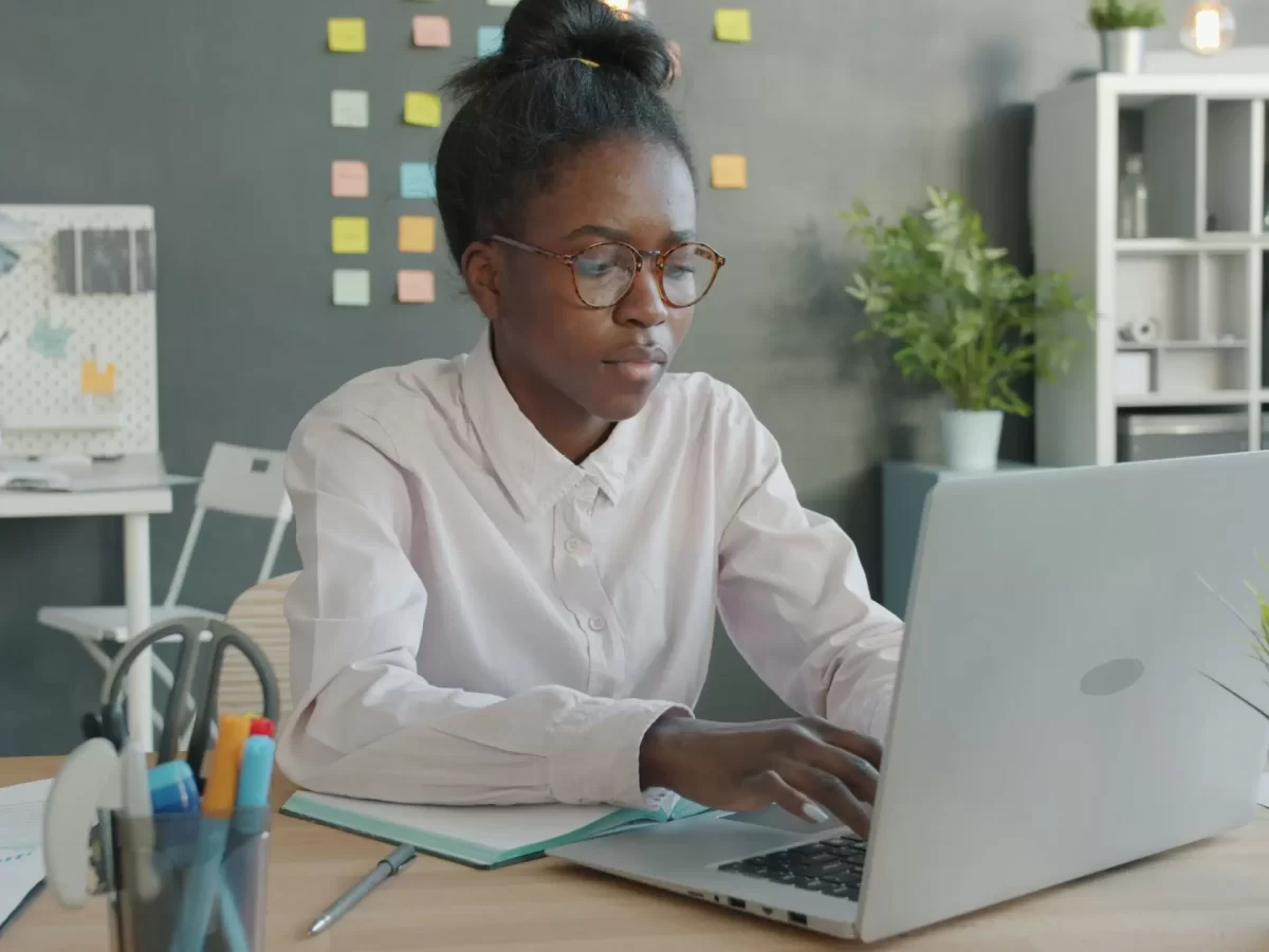 Person wearing glasses works on a laptop at a desk in a modern office with sticky notes on the wall and office supplies nearby. - PsychDiscouse