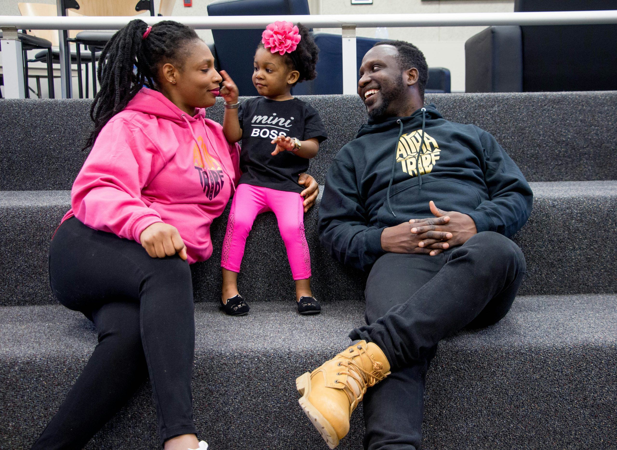 A woman, a man, and a young girl sit together on carpeted stairs, smiling and interacting. The child wears a “mini boss” shirt and a pink flower headband. - PsychDiscouse