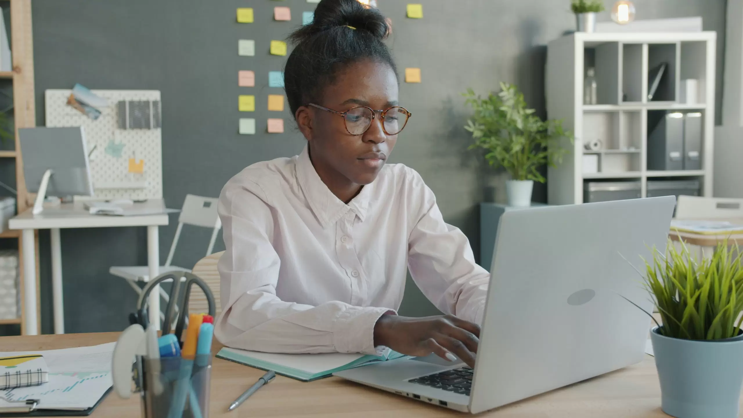 Person wearing glasses works on a laptop at a desk in a modern office with sticky notes on the wall and office supplies nearby. - PsychDiscouse